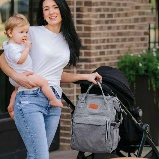 Maman tenant son bébé et poussant une poussette avec un sac à dos à langer gris accroché.